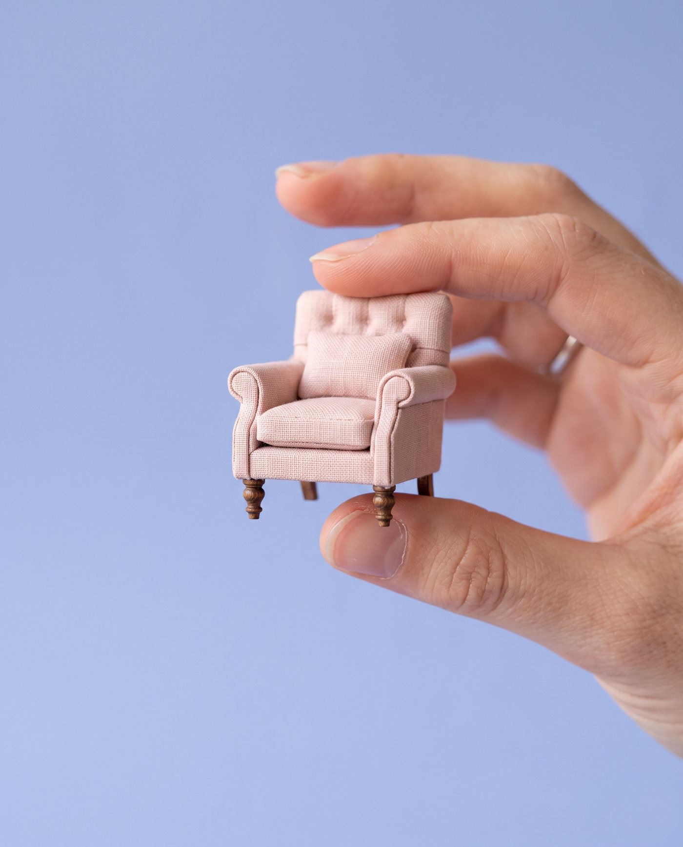 A hand holding a tiny blush pink 1:12 scale armchair between thumb and forefinger against a periwinkle background